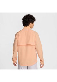 Camisa de manga larga de color melocotón con un diseño de espalda en capas, hecha de una tela ligera y texturada, que presenta un escote redondo y mangas ajustadas.