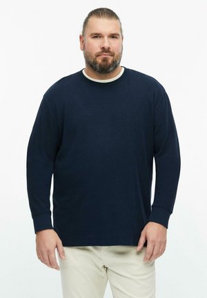 Homme avec barbe et cheveux attachés porte une chemise à manches longues bleu marine et un pantalon beige, debout devant un fond blanc uni.