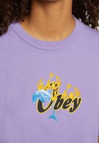 Close-up van een paars shirt met de grafische tekst "Obey" in zwarte en gele vlammen, en een blauwe dolfijn verwerkt in het ontwerp.