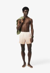 Pantaloni da bagno da uomo in tessuto pesca chiaro con cintura elastica, indossati a piedi nudi, con un asciugamano verde appoggiato su una spalla.