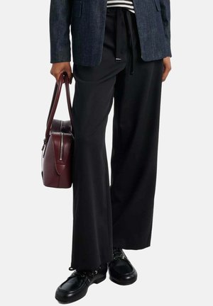 Persoon die zwarte wijde broek, zwarte schoenen, grijze blazer, gestreept overhemd draagt, en een donkerbruine leren handtas naast zich houdt.