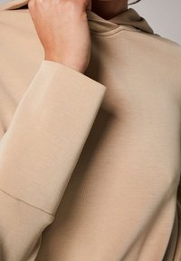 Närbild av en person som justerar den beige kragen på ett slätt, långärmat plagg med manschetter.