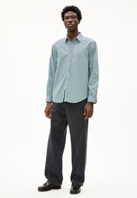 Lichtblauwe overhemdaad met een kraag, lange mouwen en donkere jeans. Model draagt zwarte instappers en staat tegen een effen witte achtergrond.