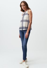Beige, ärmelloses Oberteil mit kariertem Muster und blauen Streifen, kombiniert mit schlanken blauen Jeans und weißen Sneakern. Glatte Stoffqualität, lässiges Design.