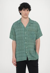 Camisa de manga curta verde com padrão, com gola recortada, apresentando designs geométricos abstratos e fechamento frontal com botões. O tecido parece leve.