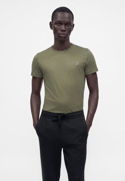 Homme portant un t-shirt kaki à manches courtes avec un petit logo zèbre et un pantalon noir à cordon, debout devant un fond blanc uni.