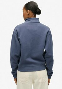 Kobieta z włosami upiętymi w kok, ubrana w niebieską bluzę z wysokim kołnierzem i jasnobezowe spodnie, widoczna od tyłu na białym tle.