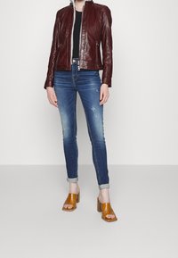 Giacca di pelle bordeaux con collo alto e chiusura con zip, abbinata a jeans aderenti blu e sandali con tacco marroni e punta aperta.