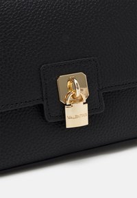 Svart texturerat lädermaterial med ett lås i gult tonläge ingraverat med "VALENTINO" på en handväsksklaff.