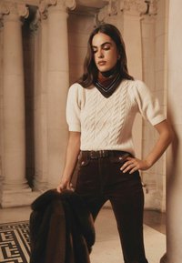 Pulover scurt cu mâneci scurte, tricotat în model cu cabluri, de culoare crem, pantaloni din catifea de culoare maro, cu talie înaltă, și o eșarfă de culoare maro închis. Modelul ține o haină din blană, pozând lângă coloane.