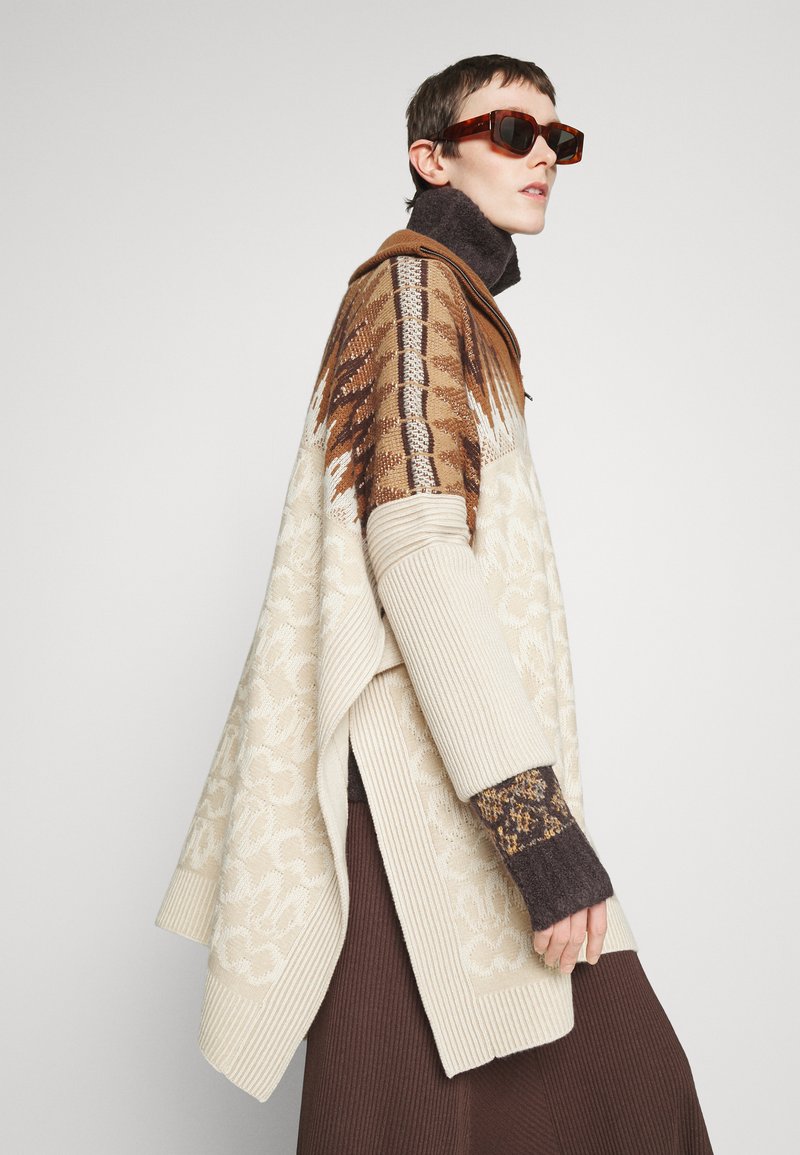 Strickponcho in Creme mit einem braunen geometrischen Muster, ausgestattet mit gerippten Bündchen und einem hohen Kragen, über einem dunkelbraunen Rock getragen.