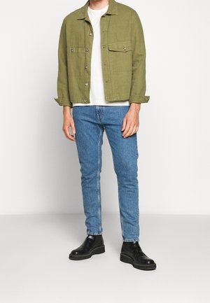 Olivgrüne Hemdjacke mit zwei Brusttaschen, kombiniert mit hellblauen Skinny-Jeans und schwarzen Stiefeletten, vor einem schlichten weißen Hintergrund.