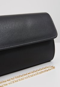Černá kabelka z umělé kůže s hladkým povrchem, zaobleným tvarem, uzavíráním na klips a zlatým řetízkem položená vedle.