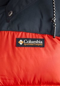 Marineblå og rød pufferjakke med Columbia-logo patch, trykknapper og et tekstureret stof med hætte, der kan justeres med snore.