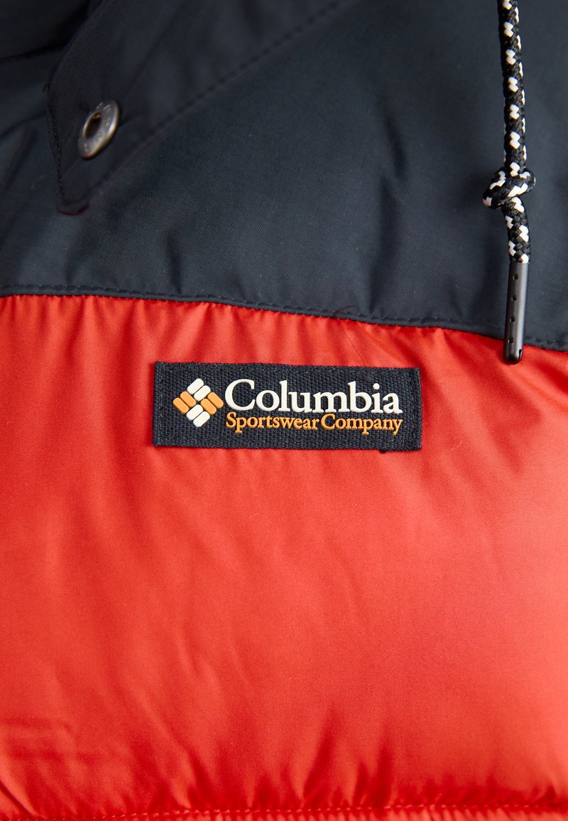 Die navyblaue und rote Steppjacke verfügt über ein Columbia-Logo-Patch, Druckknöpfe und einen strukturierten Stoff mit einer Kordelzugkapuze.