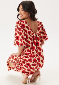 Robe à imprimé floral en crème avec des coquelicots rouges, dos en scoop, manches courtes bouffantes, détail à nouer au dos et sandales à talons blocs transparentes.