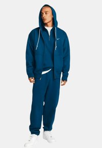 Blå zip-up hoodie med vita dragskon, kombinerad med matchande sweatpants och vita sneakers. Materialet ser mjukt ut och har en avslappnad passform.