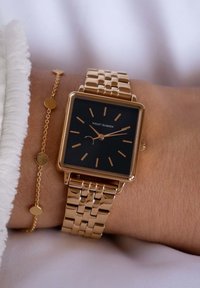 Goudkleurig vierkant horloge met een zwarte wijzerplaat, dunne uuraanduidingen en een schakelsbandontwerp. Heeft een minimalistische stijl en een zijdeachtige witte textuur in de nabijheid.
