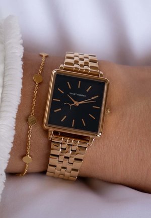 Montre carrée ton or avec un cadran noir, des marqueurs d'heure fins et un bracelet à maillons. Présente un style minimaliste et une texture blanche soyeuse à proximité.