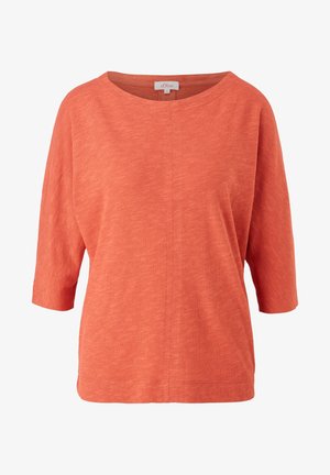Kvinde iført opkørte beige cargobukser, hvide sneakers og en løs orange t-shirt med trekvartærmer, stående mod en ensfarvet baggrund.