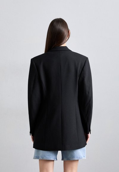 Black tailored blazer with structured shoulders, single vent at the back, worn over light blue denim shorts. Smooth fabric texture.