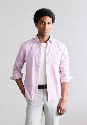 Νέος άνδρας με κοντά σγουρά μαλλιά, φοράει ένα ανοιχτό ροζ πουκάμισο πάνω από ένα λευκό μπλουζάκι, ανοιχτό γκρι παντελόνια και μαύρη ζώνη, στέκεται χαλαρός.