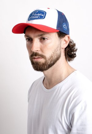 Red and white trucker cap with mesh sides, featuring a blue logo patch on the front. Worn by a man with medium-length wavy hair.