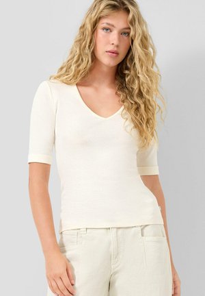 Junge Frau mit langen, lockigen blonden Haaren, die ein eng anliegendes cremefarbenes V-Ausschnitt-Oberteil mit Ellbogen-langen Ärmeln und cremefarbene Hose vor neutralem Hintergrund trägt.