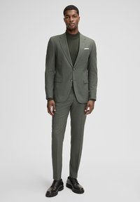 Abito due pezzi in tessuto verde oliva, vestibilità sartoriale, con giacca monopetto con rever a bavero e pantaloni slim. Indossato con un dolcevita abbinato. Scarpe eleganti nere.
