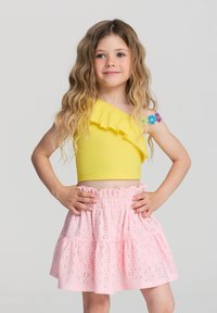 Ragazza giovane con lunghi capelli mossi, che indossa un top giallo a spalla unica e una gonna rosa a pois, in piedi con le mani sui fianchi su uno sfondo semplice.