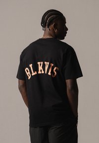 Camiseta de algodón negra con un gráfico destacado de "BLKVIS" en un color melocotón claro texturizado en la parte superior de la espalda. Mangas cortas, corte holgado.