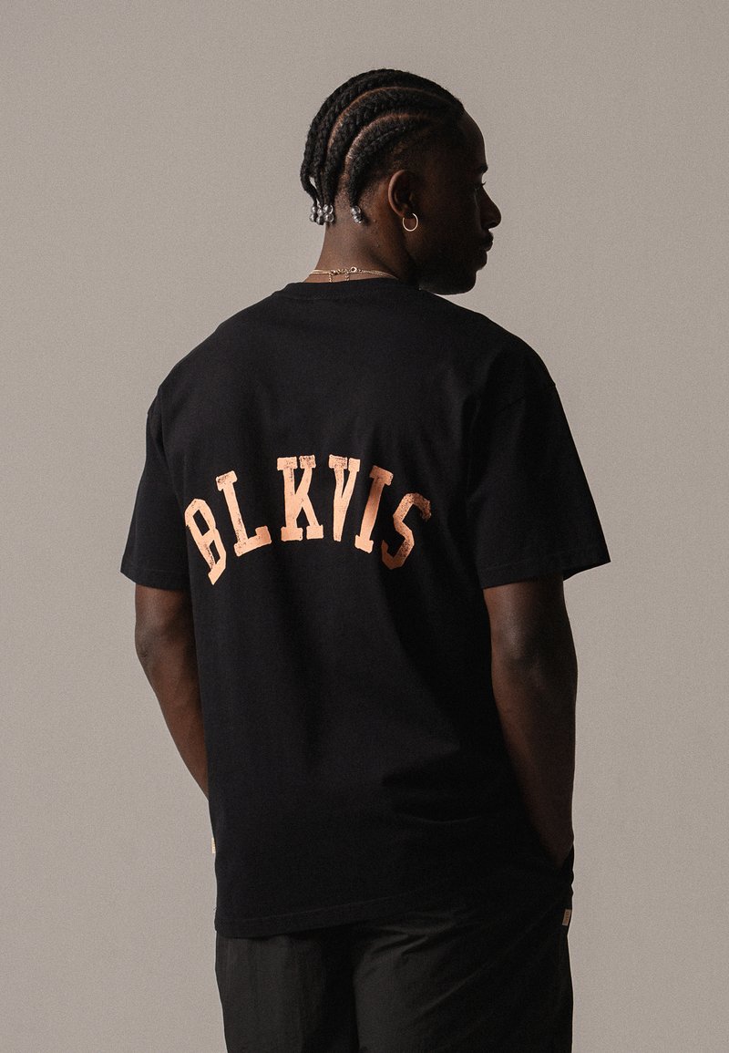 Camiseta de algodón negra con un gráfico destacado de "BLKVIS" en un color melocotón claro texturizado en la parte superior de la espalda. Mangas cortas, corte holgado.