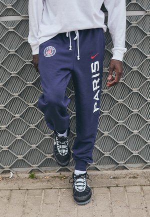 Personne portant un pantalon de survêtement Paris Saint-Germain bleu marine et des baskets noires et blanches, debout contre un mur en métal texturé.