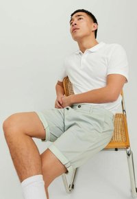 Witte poloshirt en lichtgroene gestreepte shorts met opgerolde mouwen. Model zittend op een houten en metalen stoel tegen een neutrale achtergrond.
