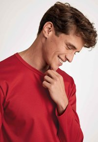 Chemise rouge à manches longues en tissu lisse, avec un col rond et des manches ajustées, portée par une personne souriante.