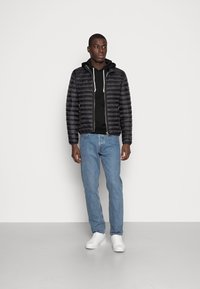Chaqueta negra acolchada con diseño acolchado horizontal, llevada sobre una sudadera negra con capucha. Jeans de mezclilla azul claro y zapatillas blancas complementan el atuendo.