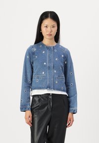 Denim jakna s okroglim izrezom, ki ima več srebrnih luknjic, sprednje žepe in skrajšan kroj, v kombinaciji z črnimi hlačami.