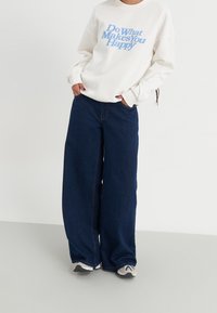 Felpa bianca con testo blu, abbinata a jeans chiari in denim scuro a vestibilità larga. I jeans presentano tasche laterali e una silhouette rilassata.