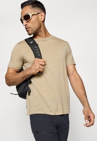Camisa de manga corta beige en tejido liviano, combinada con una mochila negra y gafas de sol. La mochila cuenta con correas de hombro acolchadas.