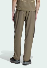 Pánske olivovo-zelené športové nohavice so voľným strihom, so bočnými pruhmi, hladkou textúrou a elastickým pásom. Kombinované s čiernymi topánkami.