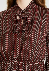Blus i röd och svart mönster med knytkrage, volangkrage och vertikalt randig design. Lättviktsmaterial med mjuk textur.