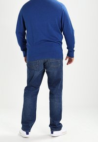 Homme debout de dos, portant un pull bleu, un jean bleu et des baskets blanches, sur un fond blanc.