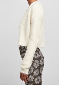 White fuzzy cropped sweater with long sleeves, featuring a soft texture. Worn with patterned brown leggings showcasing white floral designs.