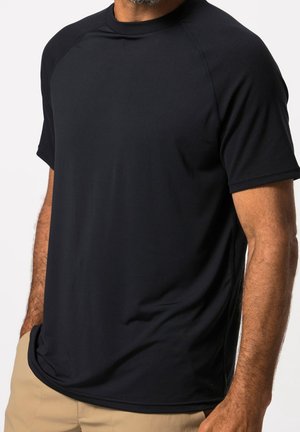 Mann trägt ein einfaches schwarzes T-Shirt mit kurzen Raglanärmeln und beiger Hose, steht vor einem weißen Hintergrund.