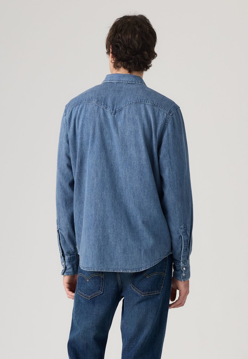Homme debout, de dos, portant une chemise en jean bleue et un jean bleu sur un fond clair uni.