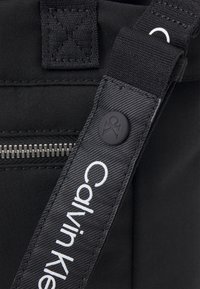 Detail černé látkové tašky s uzávěrem na zip, texturovaným popruhem s brandingem "Calvin Klein" a kulatým černým tlačítkem jako akcentem.