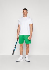 Maglietta atletica bianca con logo verde Lacoste, abbinata a pantaloni corti in rete verdi, sneakers bianche e in mano una racchetta da tennis e una pallina.