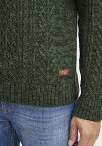 Sötétzöld kötött pulóver, texturált kábel mintával. Bordás mandzsettával és alsó szegéllyel rendelkezik. Oldal�án lévő bőr címke felirattal. Kék farmernadrággal viselve.