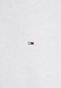 Witte gebreide trui met een V-hals en een klein geborduurd logo in rood, marineblauw en wit aan de linkerkant.