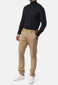 Chino beige dengan potongan lurus, ritsleting, kantong depan, dan kancing cokelat. Dipadukan dengan turtle neck lengan panjang hitam dan sepatu sneakers putih.